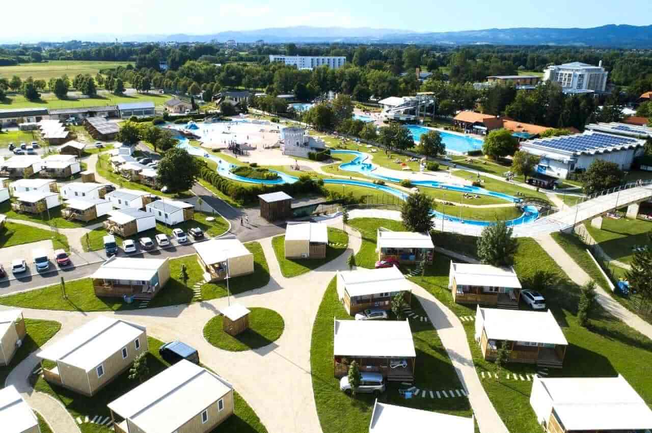 Aerial view of a holiday village with white cabins, winding paths, and a large community pool complex.