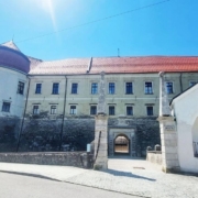Historic stone fortress with a curved tower, red roof, and arched gate across a cobblestone street.