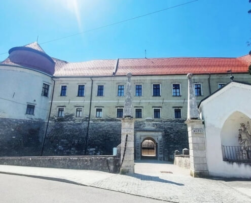 Historic stone fortress with a curved tower, red roof, and arched gate across a cobblestone street.