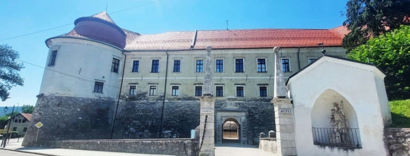 Historic stone fortress with a curved tower, red roof, and arched gate across a cobblestone street.