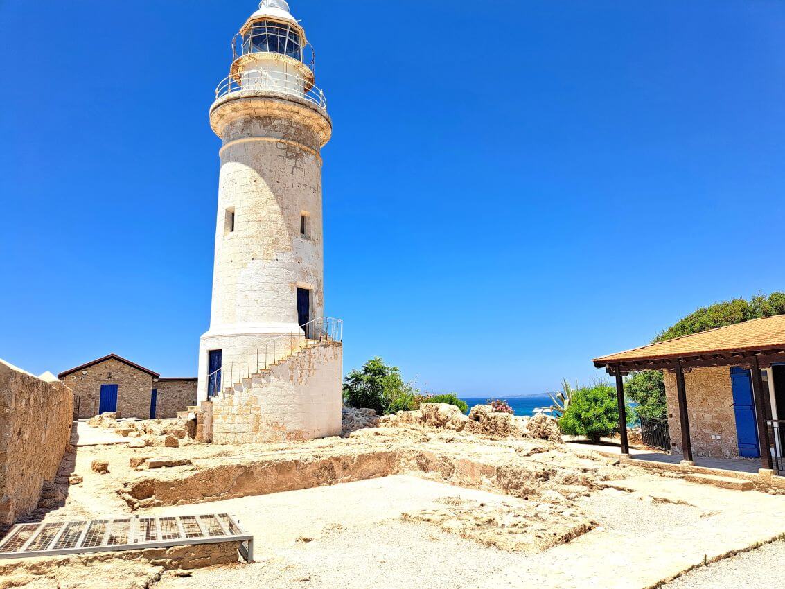 Tall white lighthouse with a spiral staircase leading to the lantern room, set against a bright blue sky near a rocky coastal area.